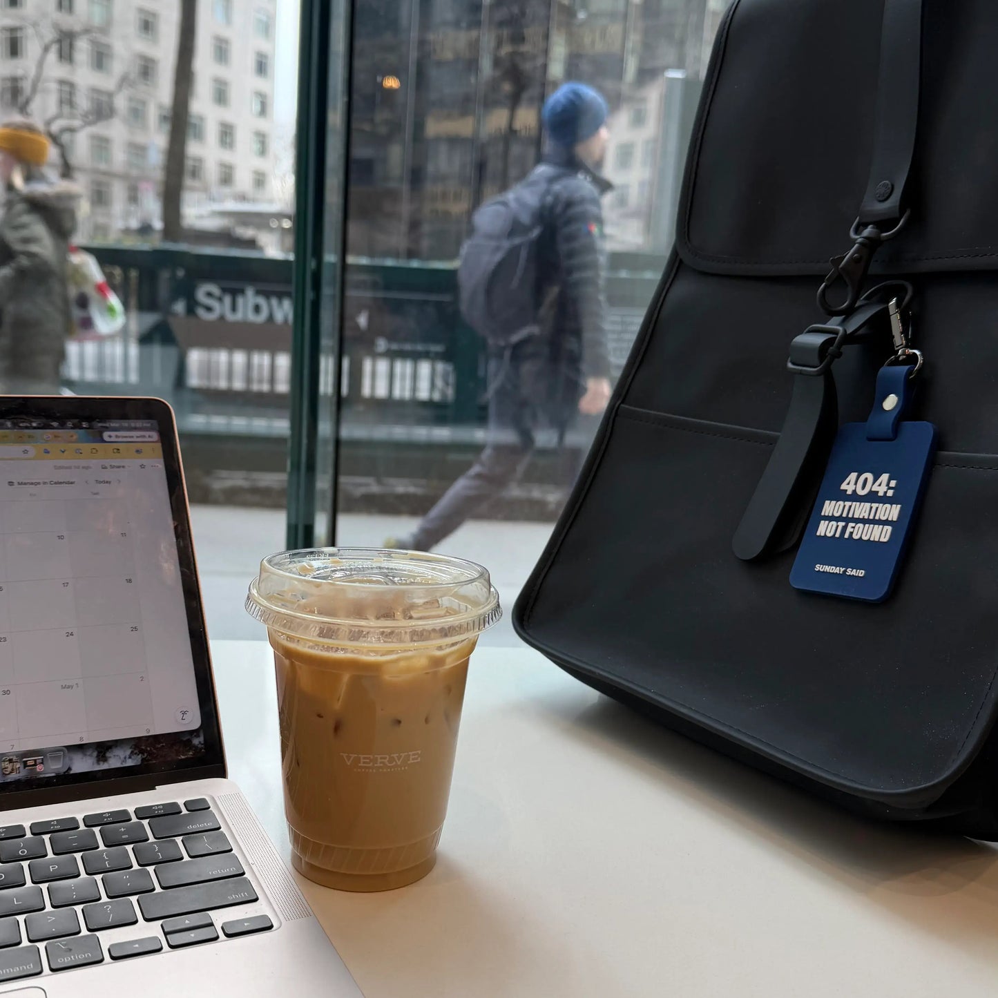 Laptop, coffee cup, and backpack on a table with a blue keychain reads 404: motivation not found