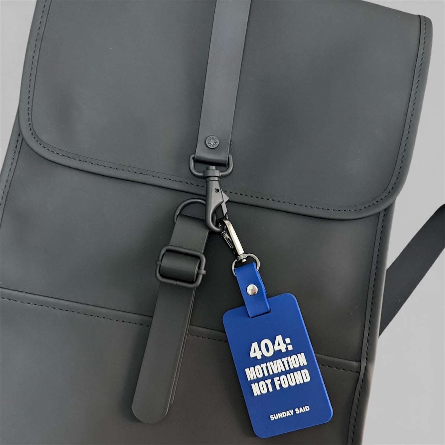 Blue 404 Motivation Not Found charm attached to a matte black minimalist backpack. Funny corporate humor bag accessory for work commute by Sunday Said.
