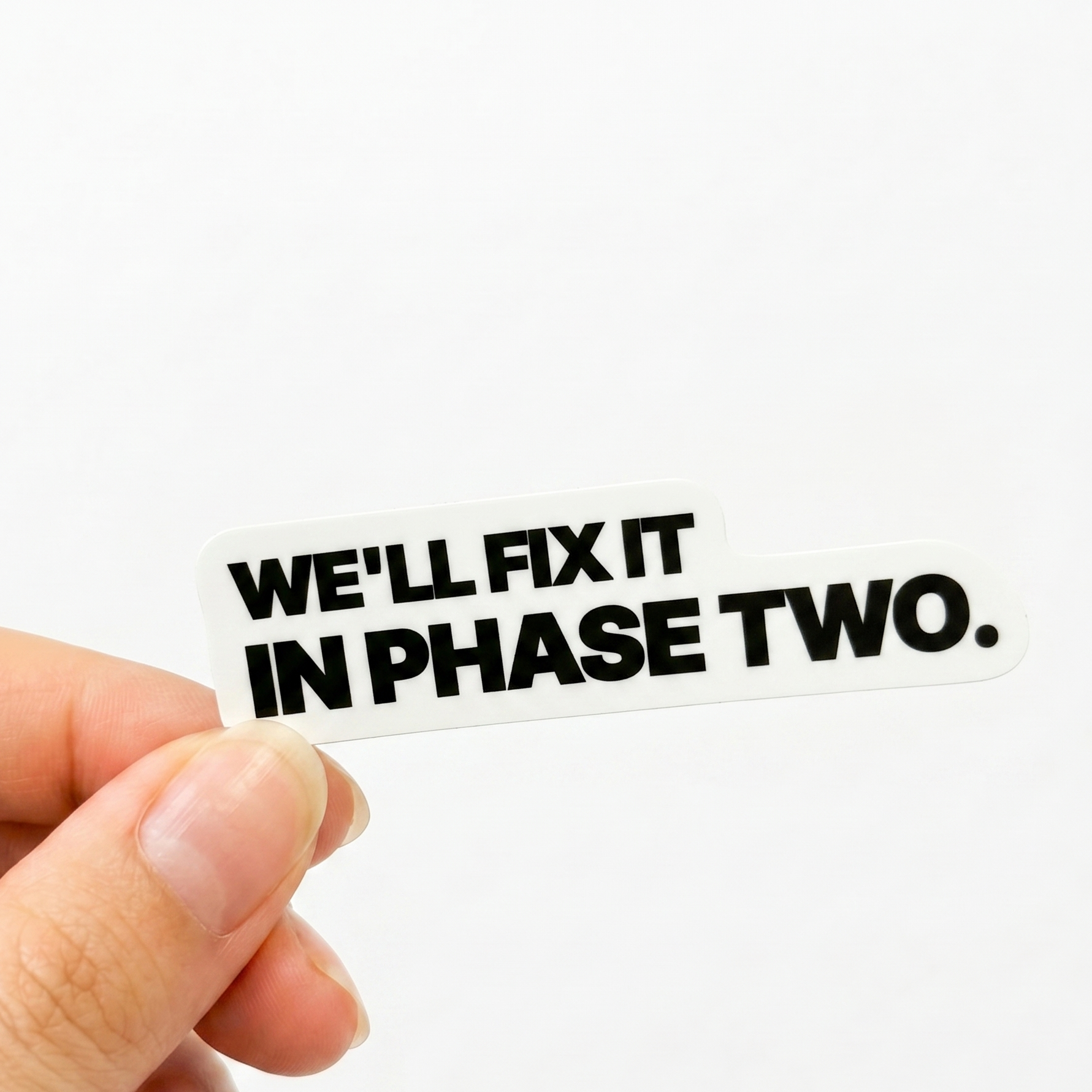 White workplace sticker with black text reading “We’ll fix it in phase two.”