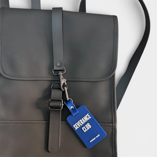 Blue Severance Club bag charm attached to a matte black minimalist backpack. Funny layoff gift and corporate resilience accessory by Sunday Said.